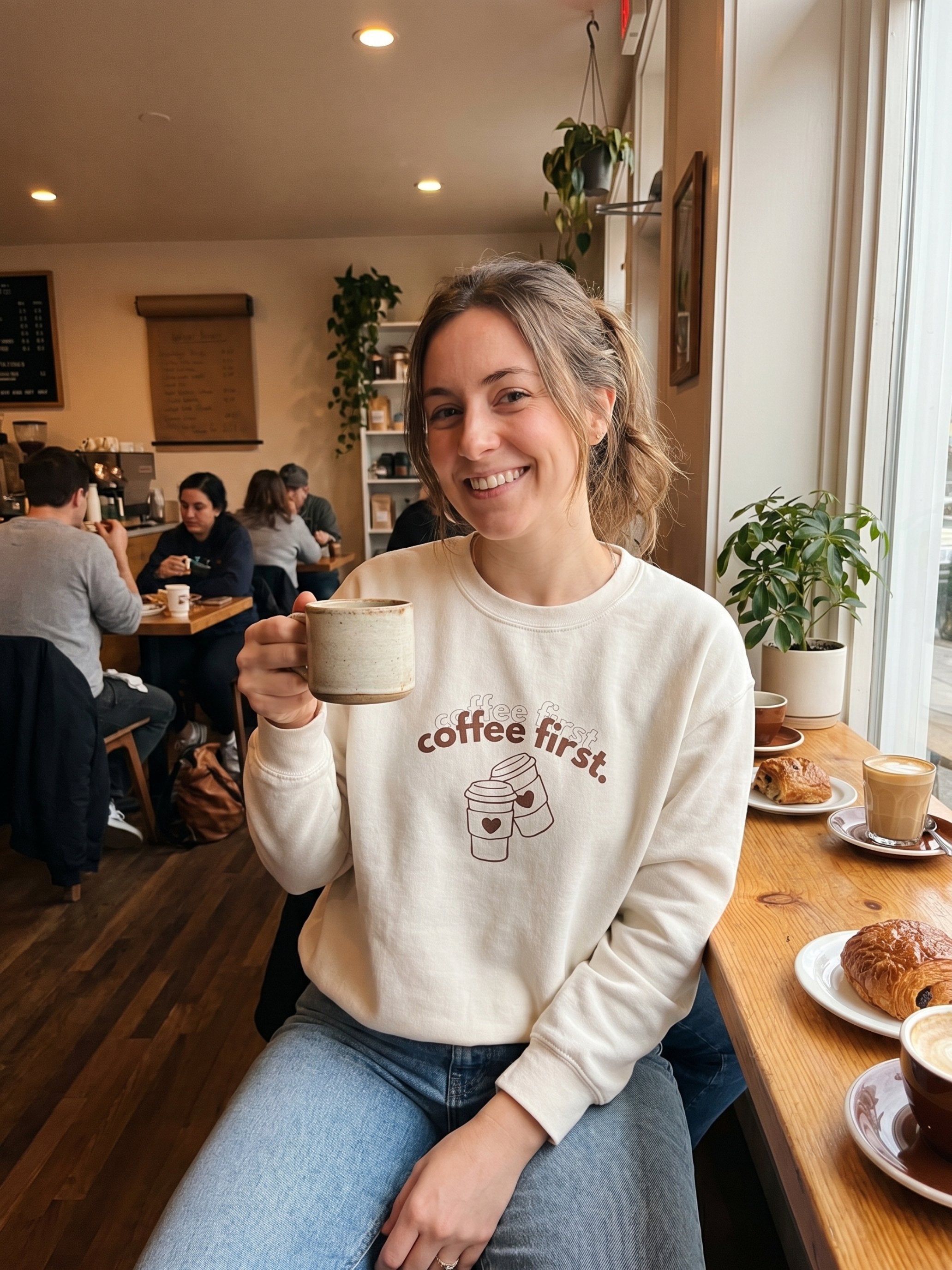 Woman in a coffee shop holding a mug with a 'coffee first' sweatshirt.