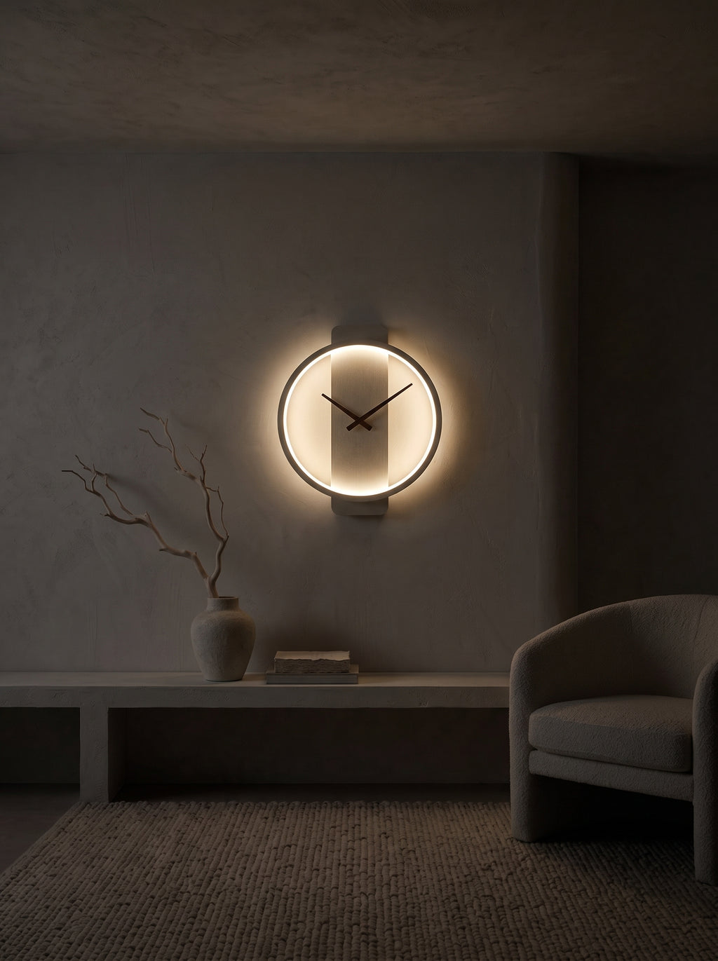 A modern circular wall clock with a glowing warm light and black hands, mounted on a textured gray wall in a dimly lit minimalist room with a bench, vase, and chair.