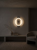 A modern circular wall clock with a glowing warm light and black hands, mounted on a textured gray wall in a dimly lit minimalist room with a bench, vase, and chair.