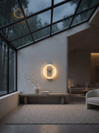 A minimalist round wall clock with a glowing warm white LED ring and black hands, mounted on a textured light gray wall in a modern, sunlit interior with a glass skylight.
