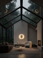 A modern living room with a large glass gable roof overlooking a rain-slicked forest. The interior features a curved gray sofa, a light beige armchair, a low glass coffee table, and a glowing circular wall clock centered on the wall.