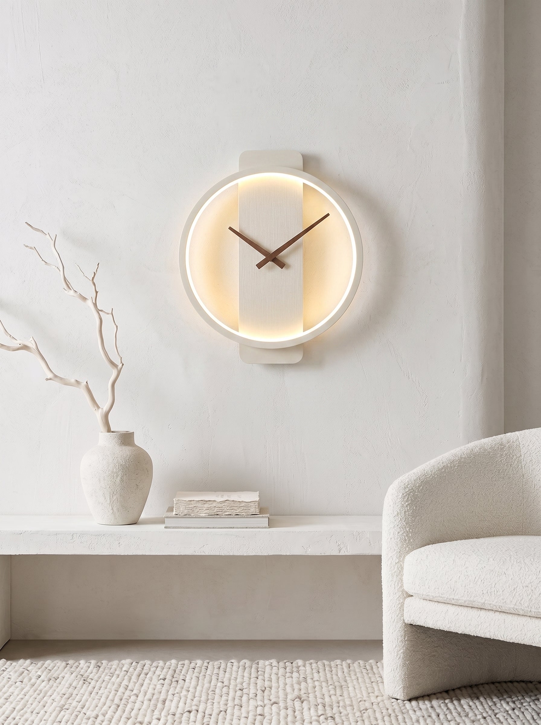 A modern circular wall clock with a warm glowing LED light ring and dark wood hands, mounted on a textured white wall above a minimalist shelf with a vase and books, next to a cream-colored armchair.