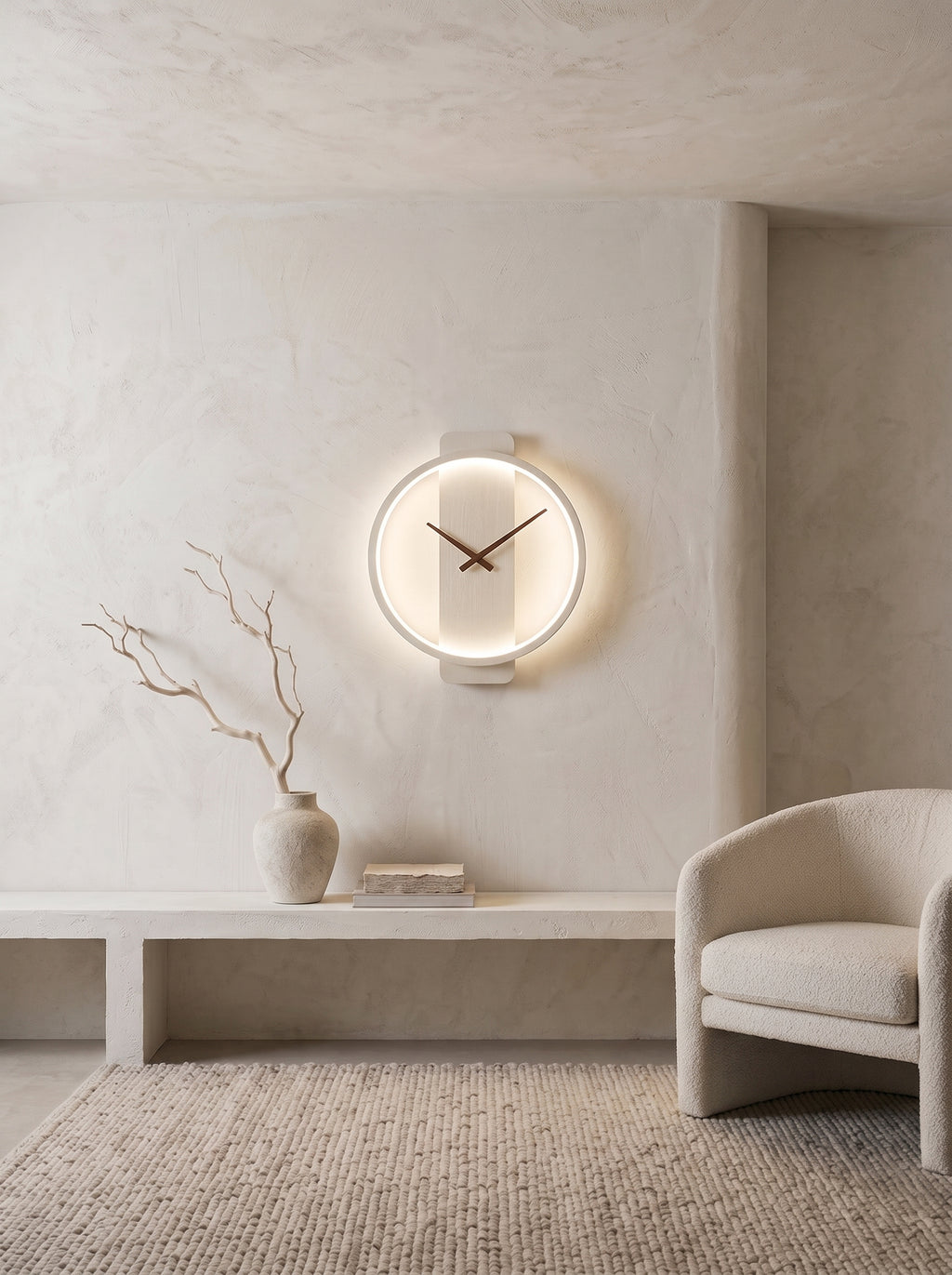 A minimalist circular wall clock with a glowing white LED ring and dark hands, mounted on a textured plaster wall in a modern, neutral-toned interior next to a light beige armchair.