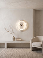 A minimalist circular wall clock with a glowing white LED ring and dark hands, mounted on a textured plaster wall in a modern, neutral-toned interior next to a light beige armchair.