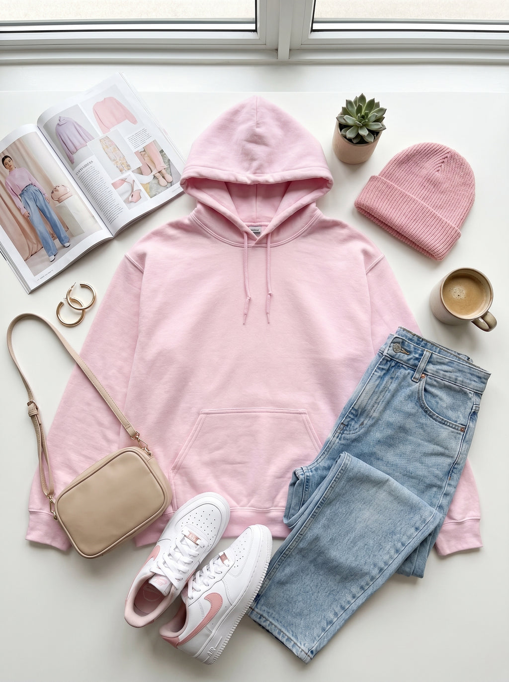 Pink hoodie, jeans, sneakers, and a beanie arranged on a light surface with a magazine and plant in the background.