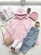Pink hoodie, jeans, sneakers, and a beanie arranged on a light surface with a magazine and plant in the background.