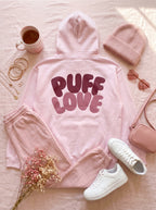 Pink 'Puff Love' hoodie with matching pants, white sneakers, a beanie, and a cup on a light pink surface.