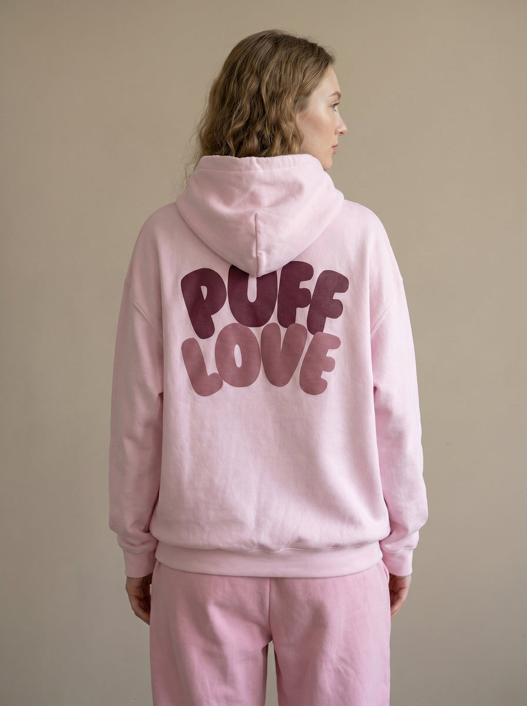 Person wearing a pink hoodie with 'PUFF LOVE' text on the back against a beige background
